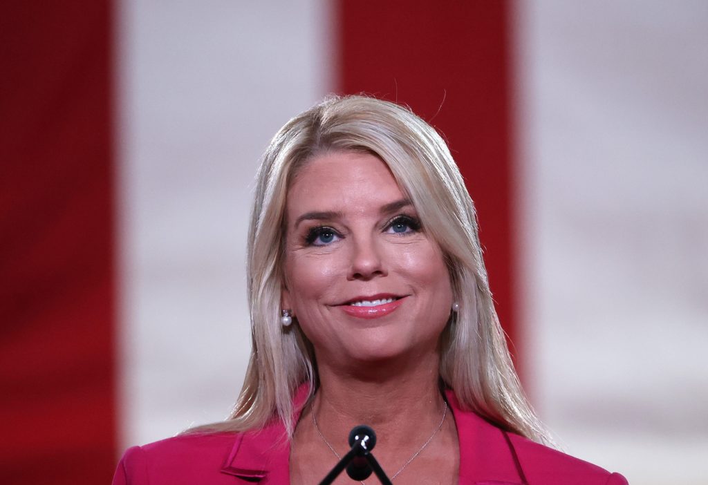 Attorney General Pam Bondi speaking at a podium during an official event, wearing a red blazer.