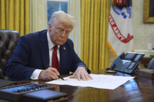 President Donald Trump signing a bill at his desk in the Oval Office, focused on the document in front of him.
